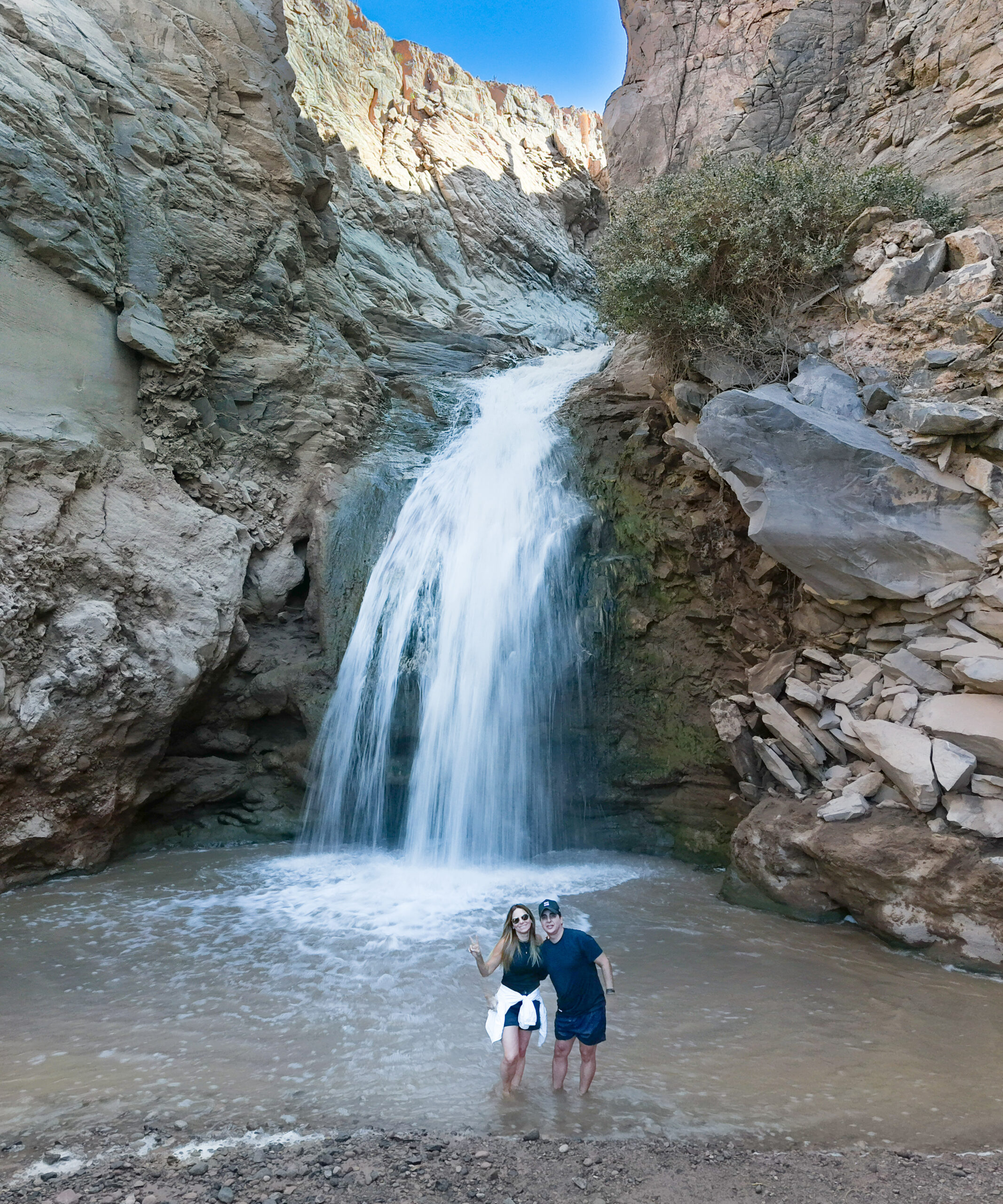 Fotografía de una pareja frente a la Cascada escondida, entre rocas, lugar en el que termina un bonito hiking cerca de San Pedro de Atacama.