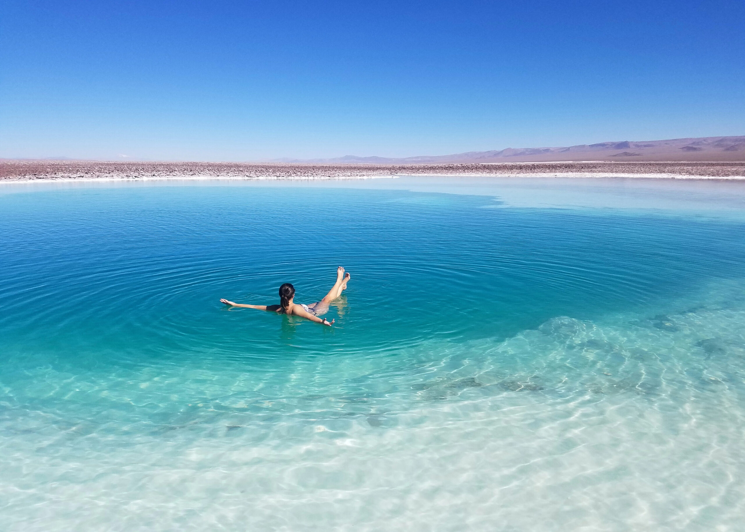 Fotografía de una chica flotando en una de las lagunas escondidas de Baltinache, rodeada de sal y con volcanes en el horizonte. En San Pedro de Atacama.